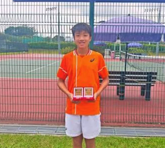 A young tennis player holding two awards beside an outdoor court